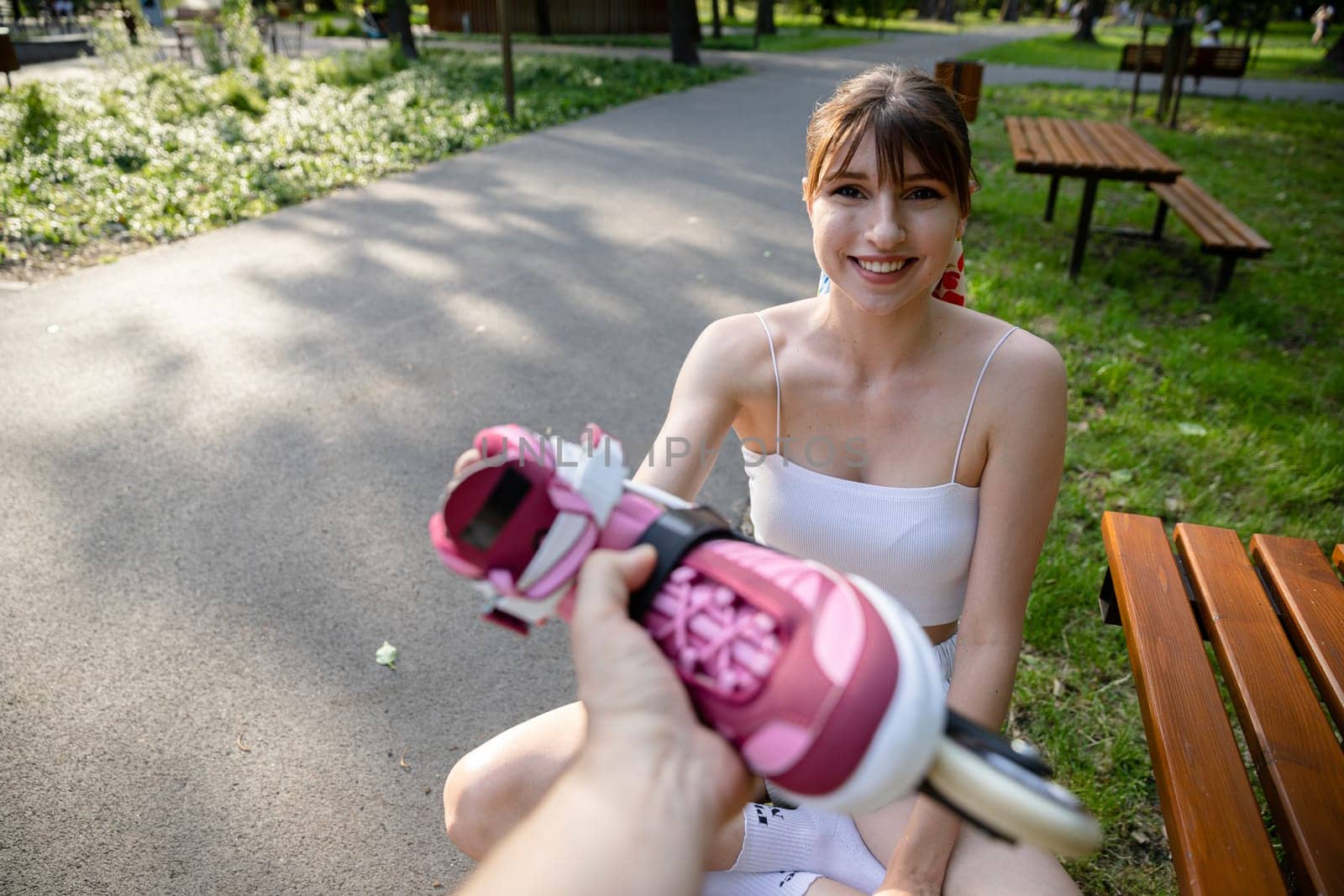 In the blurred foreground, a hand passing a rollerblade to a girl sitting on a bench. Smiling young girl combed in ponytail. White matching sports outfit.
