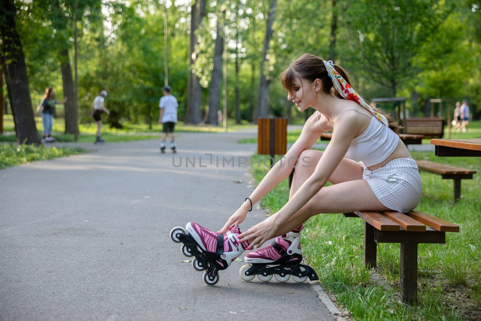 A woman improves the fastening of rollerblades while sitting on a bench. A girl puts on rollerblading sports equipment. In the distance, blurry figures of people playing sports and walking down an alley.