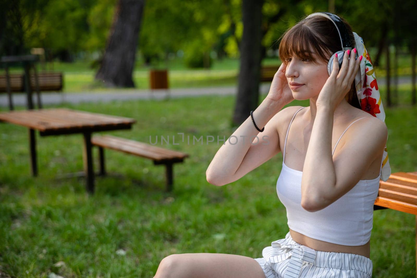 The girl puts headphones on her head. The woman is styled in a ponytail tied with a colorful scarf. She has pink fingernails. A young person sits in a park, surrounded by trees and greenery.