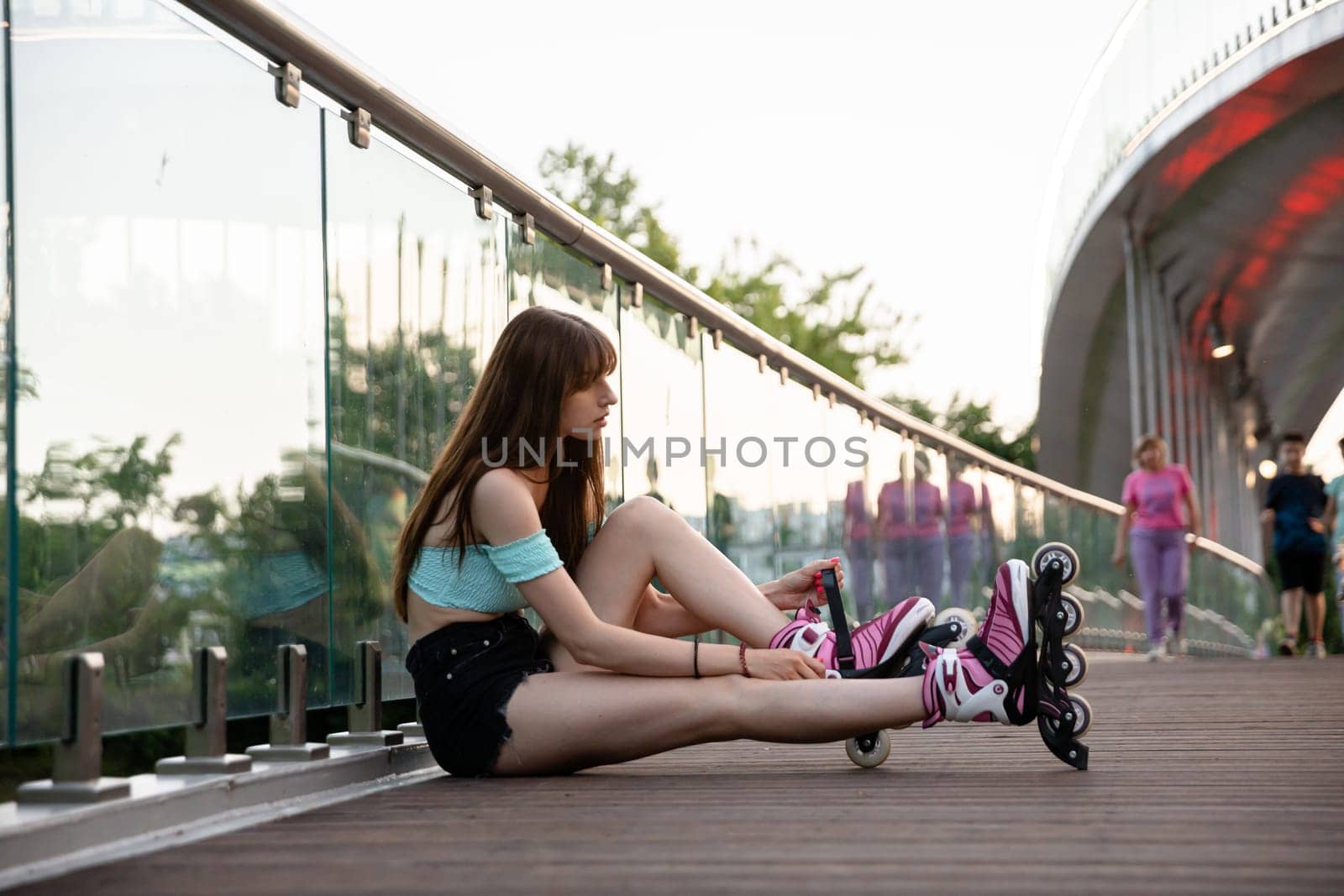 A long-haired girl sits on a bridge and puts on rollerskates. She has a glass railing behind her back. In the background you can see blurred figures of people walking.