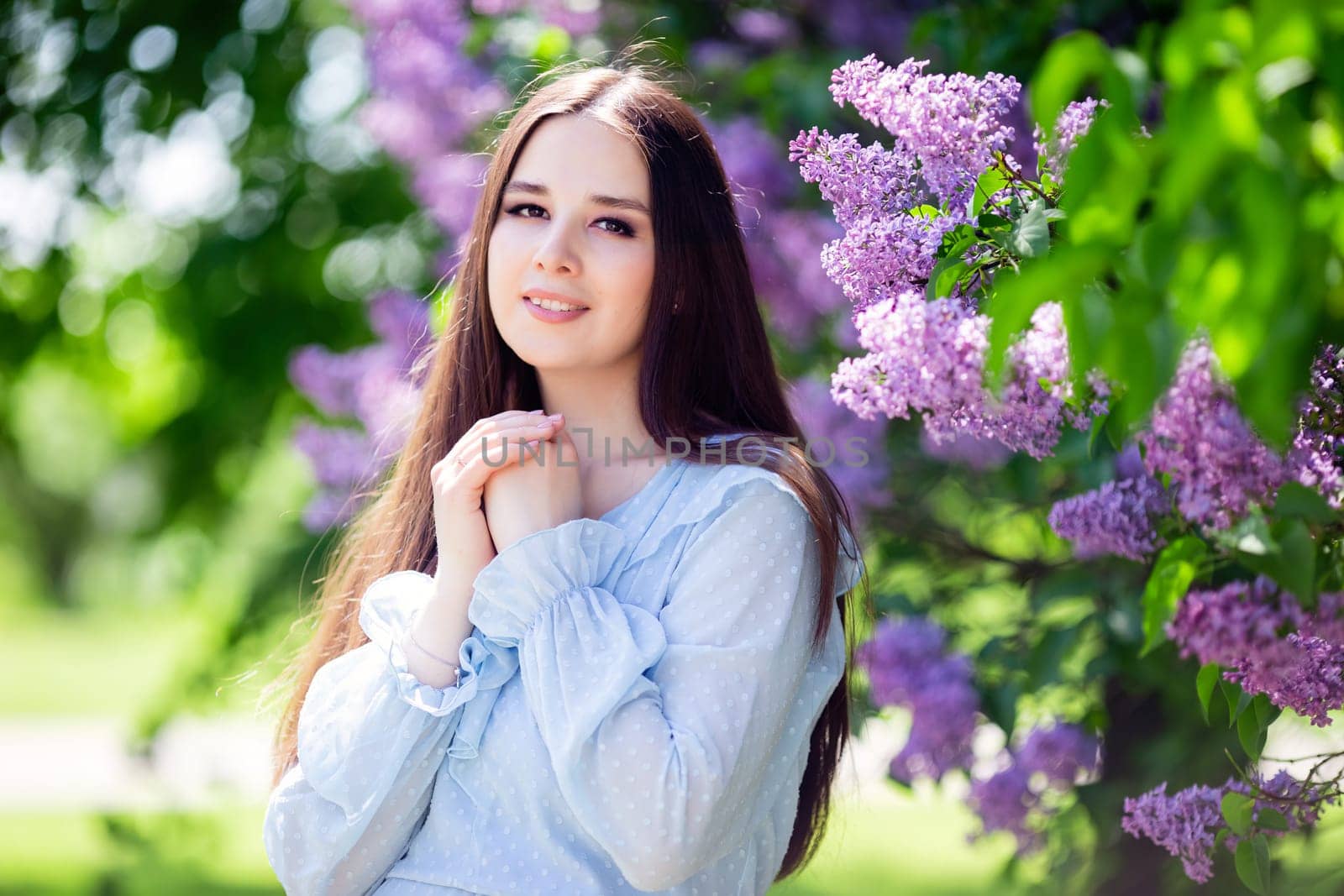 Pretty smiling brunette girl with long hair stands with lilac flowers, in the garden, in sunny day, look at camera. Close up. copy space