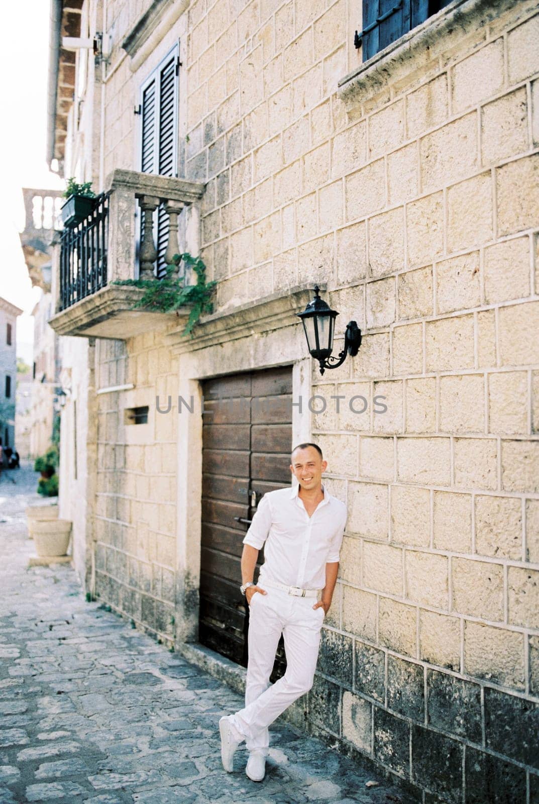 Smiling man in a white shirt stands leaning against an old stone house. High quality photo