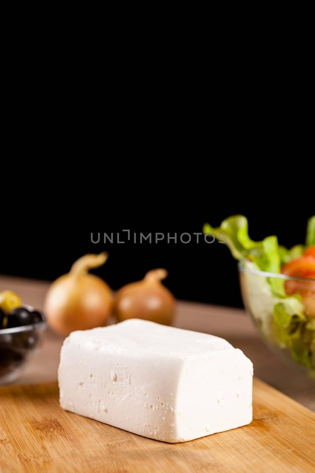 Close up on feta cheese lying on wooden board next to onions, fresh salad and glass bowl of olives on black background