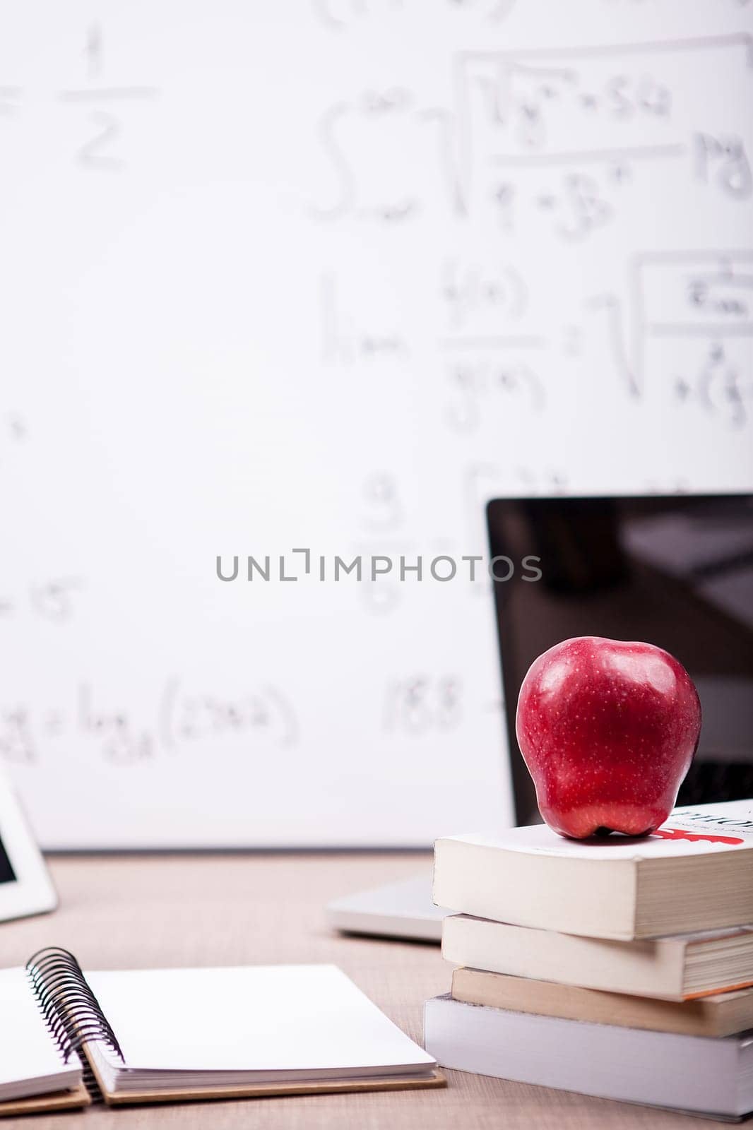 Red apple on pile of books, notebook and pencils on table with a blurred white board in the back. School concept