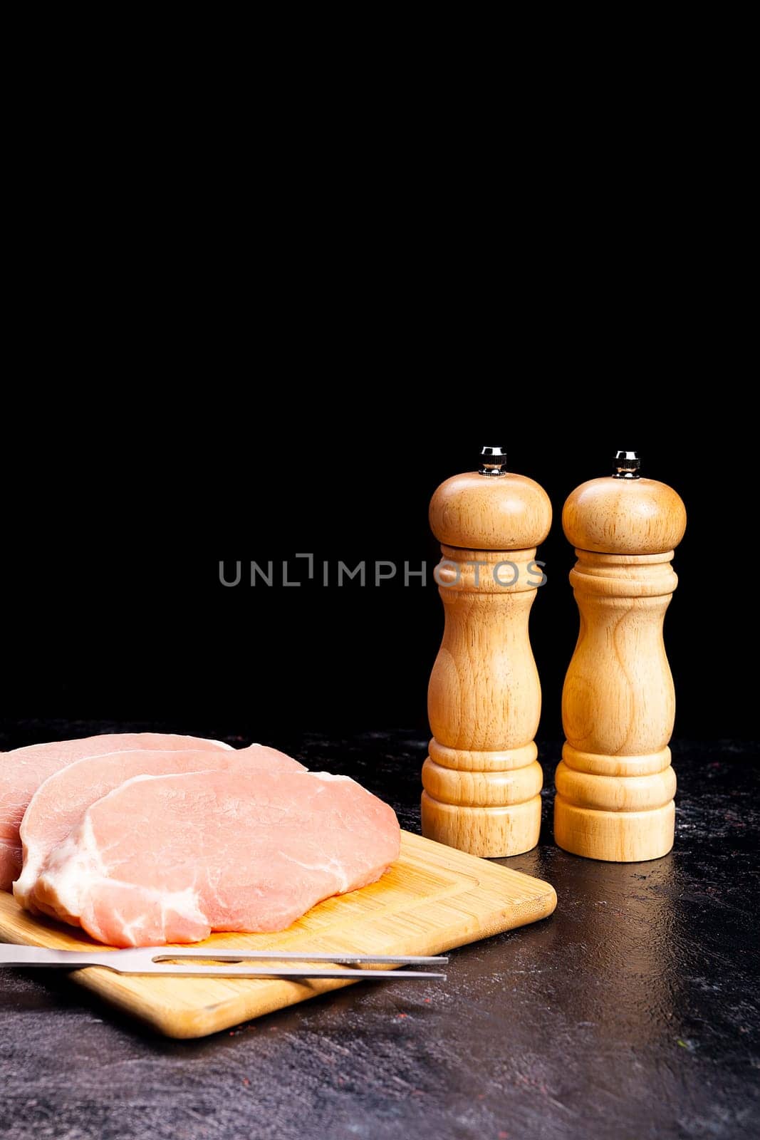 Close up on pieces of raw steaks next tot two salt and pepper shakers on black wooden background