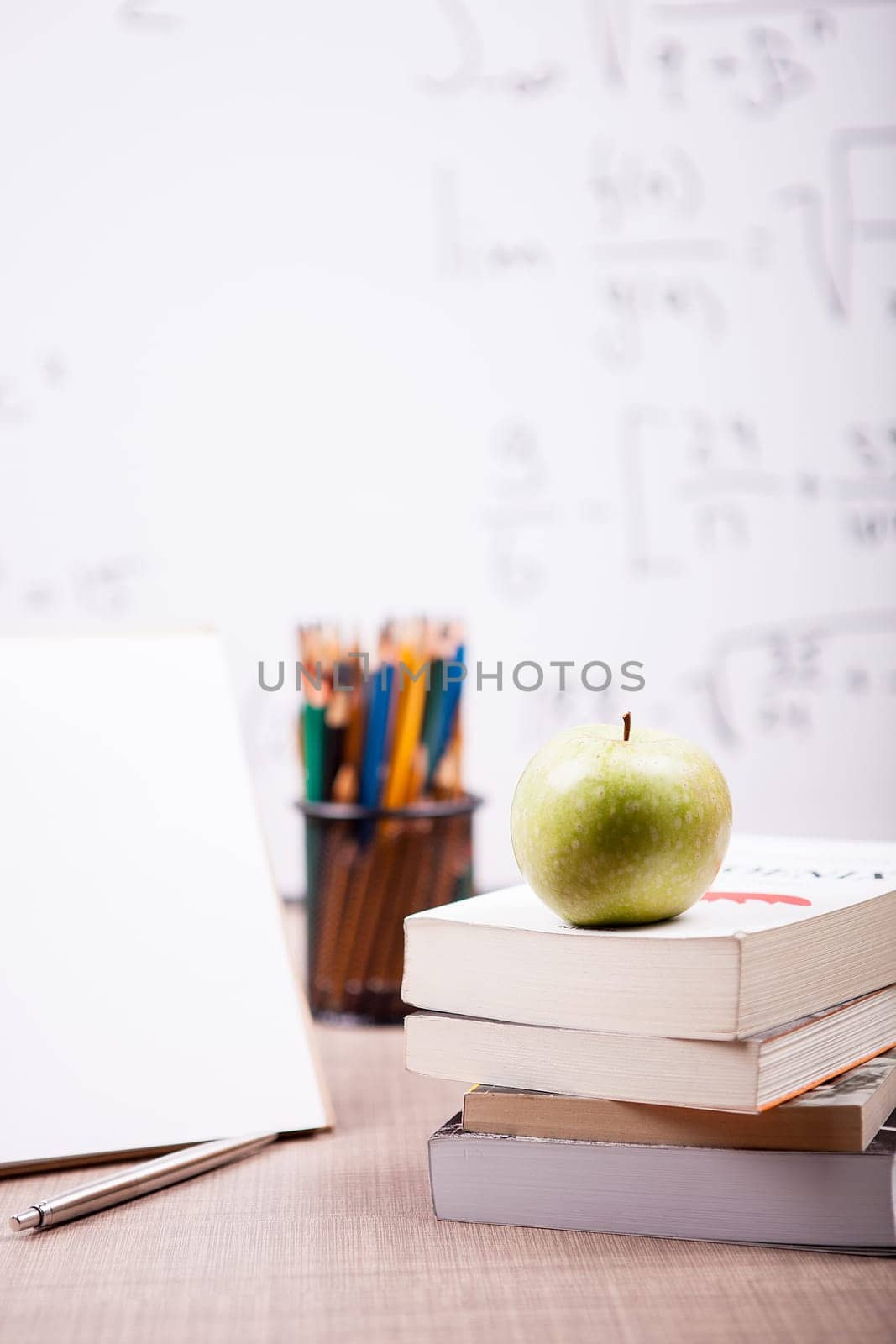 Green apple on pile of books next to a notebook and pencils on table with a blurred white board in the back. School concept