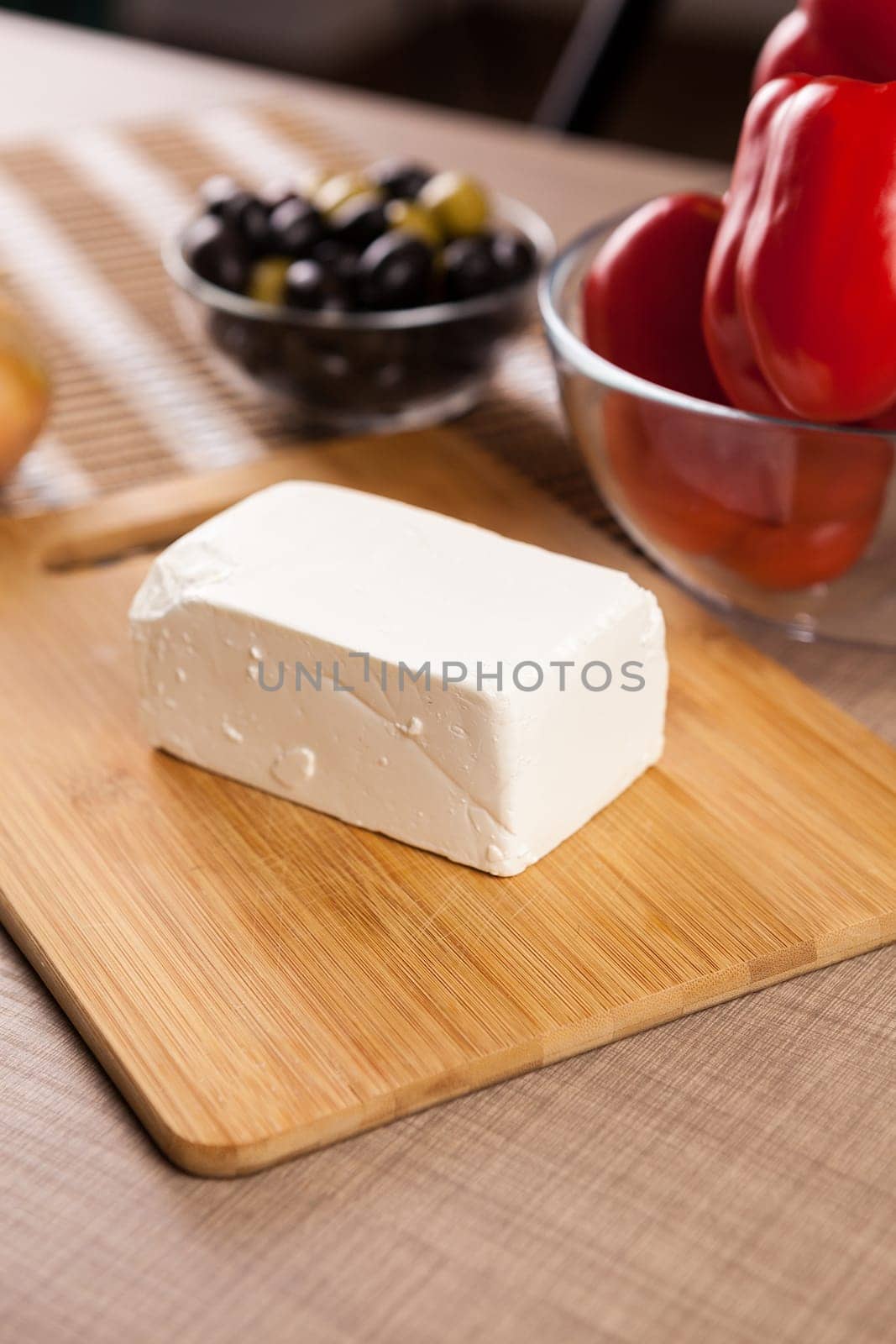 Feta chees on wooden board on a table next to a glass bowl of sweet red pepper and olives