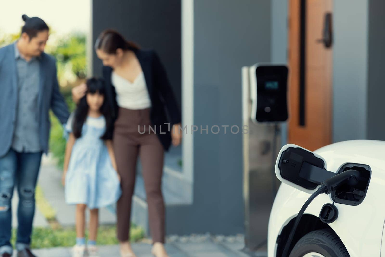 Focus closeup electric vehicle recharging battery from home electric charging station with blurred family in background. Renewable clean energy car for progressive eco awareness lifestyle concept.