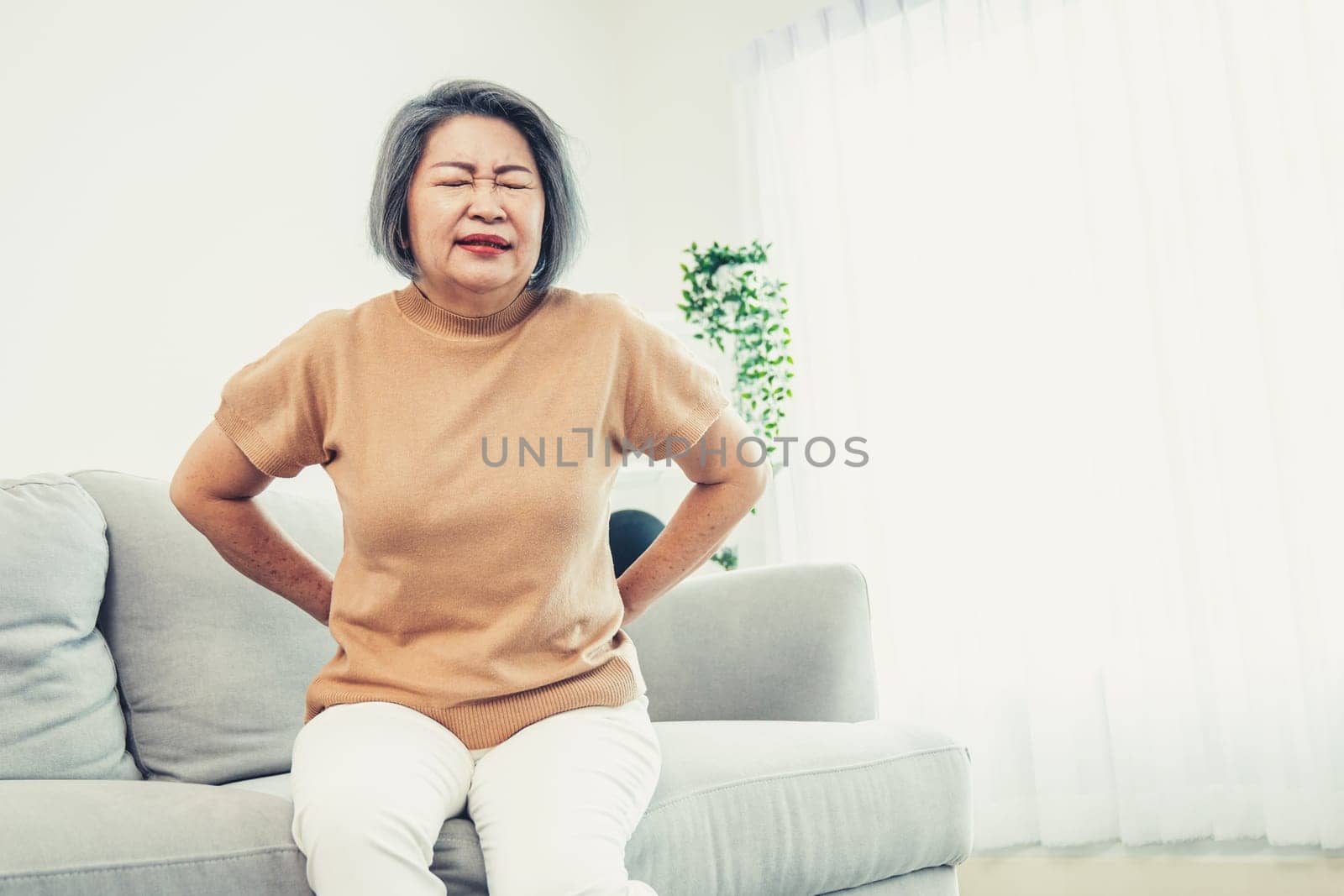 An agonizing senior woman experiencing back pain while sitting on her sofa at home and holding her back.