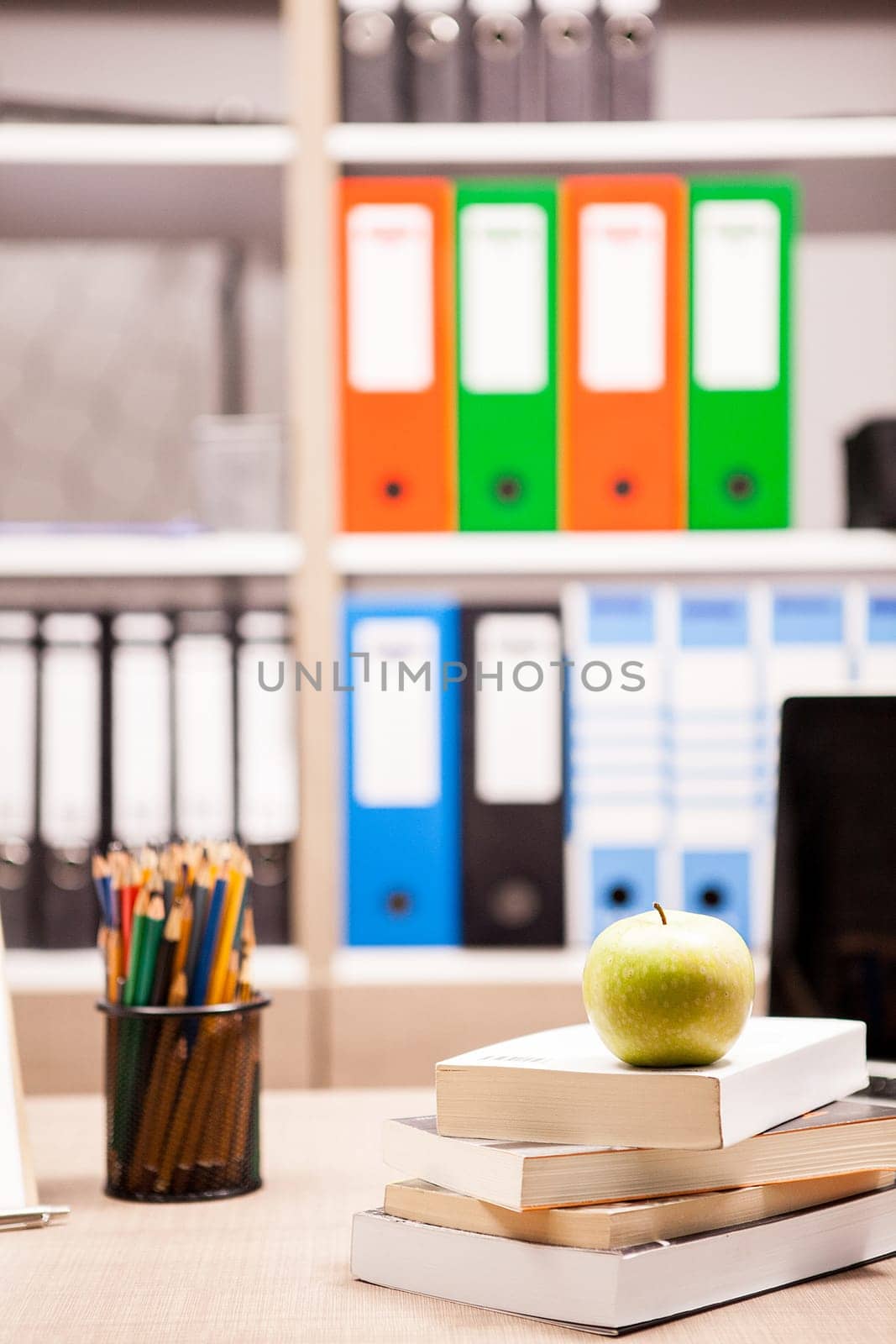 Green apple on pile of books next to a notebook and pencils on table with a blurred white board in the back. School concept