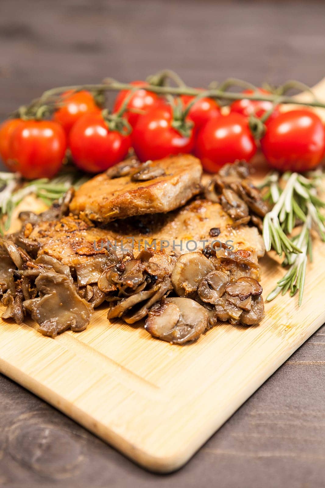 Close up on wooden board with grilled mushrooms and pork steak next to cherry tomatoes on a wooden table