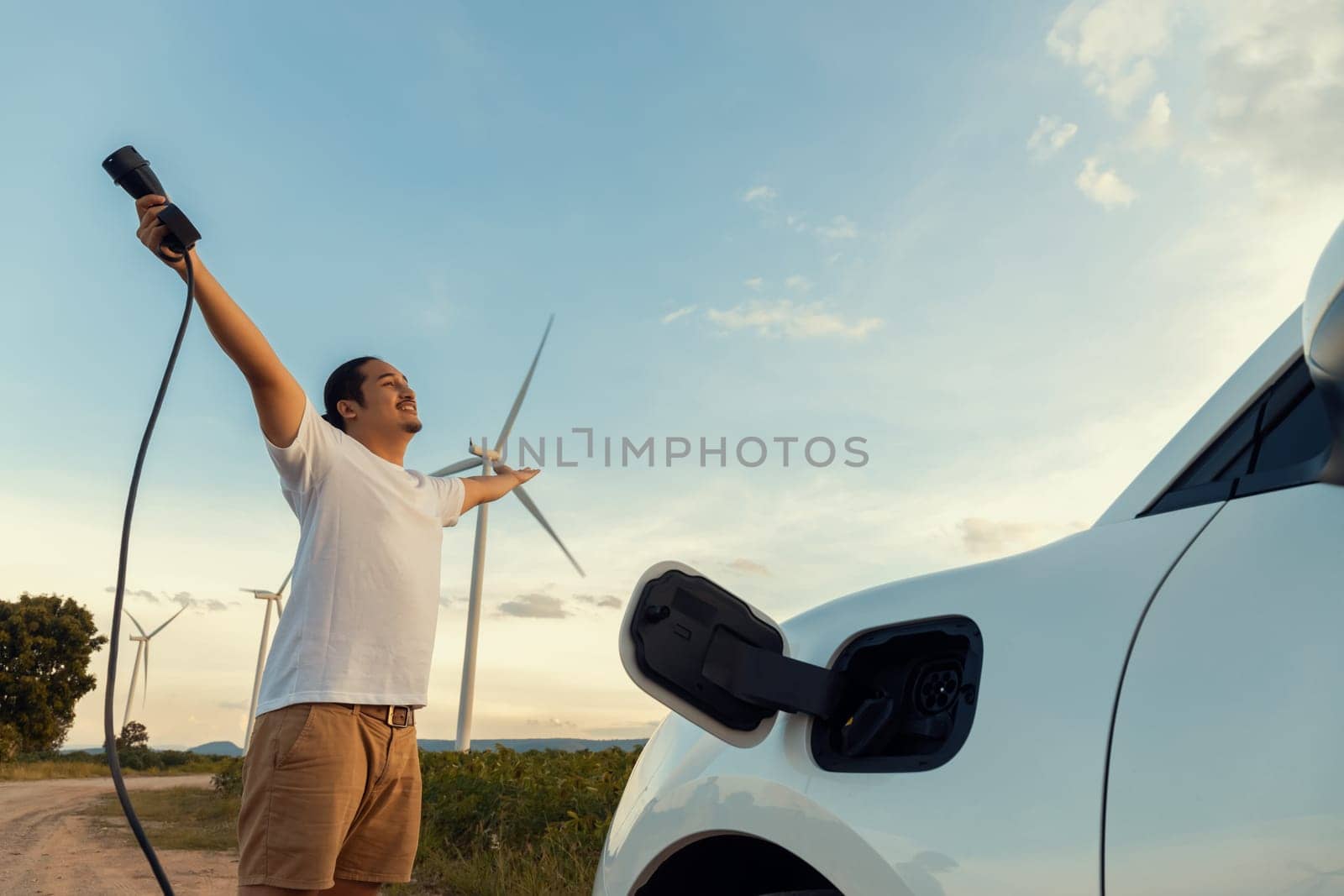 Progressive man with his EV car and wind turbine as concept of renewable energy. by biancoblue