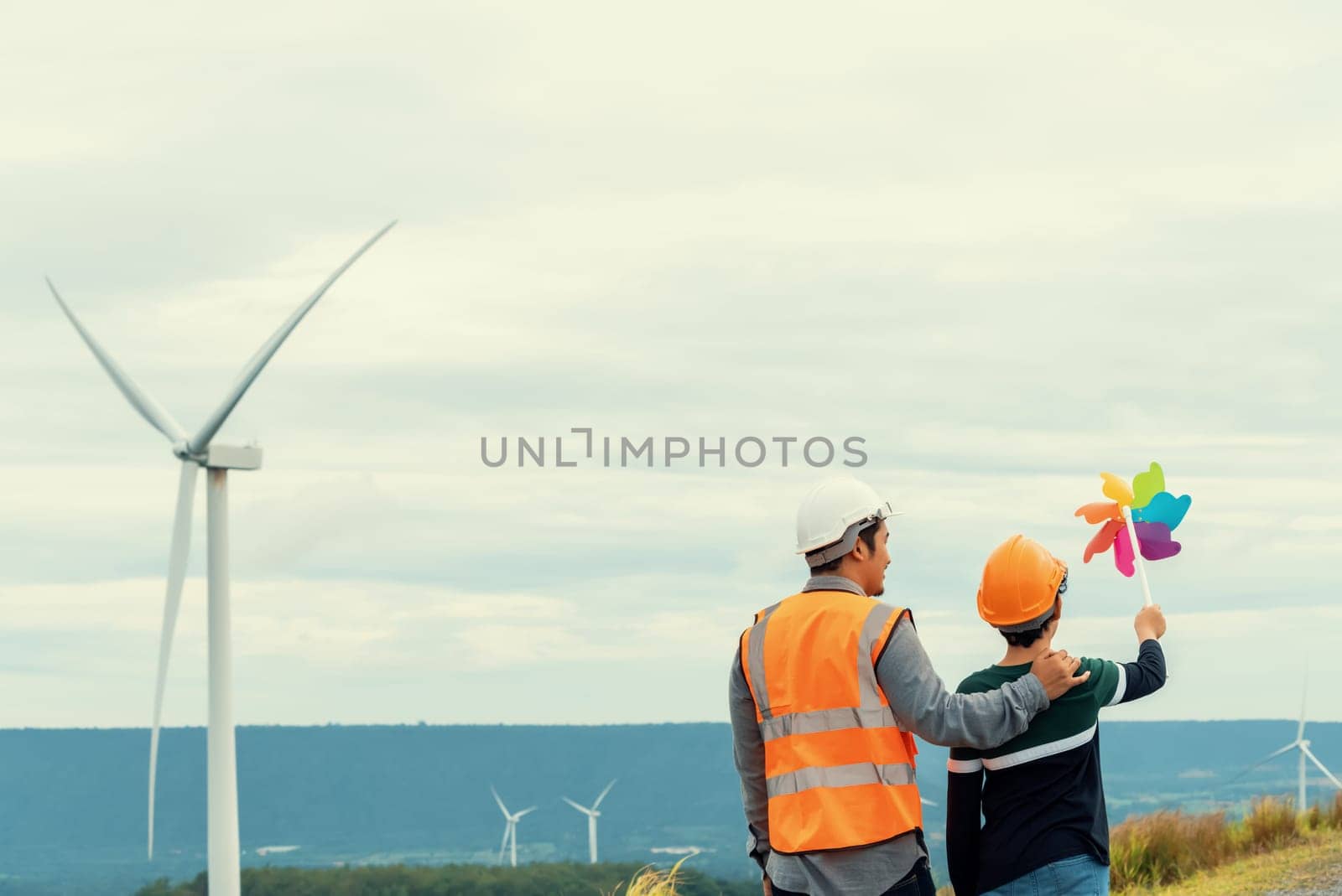 Engineer with his son holding windmill toy on a wind farm atop a hill or mountain. Progressive ideal for the future production of renewable, sustainable energy. Energy generated from wind turbine.