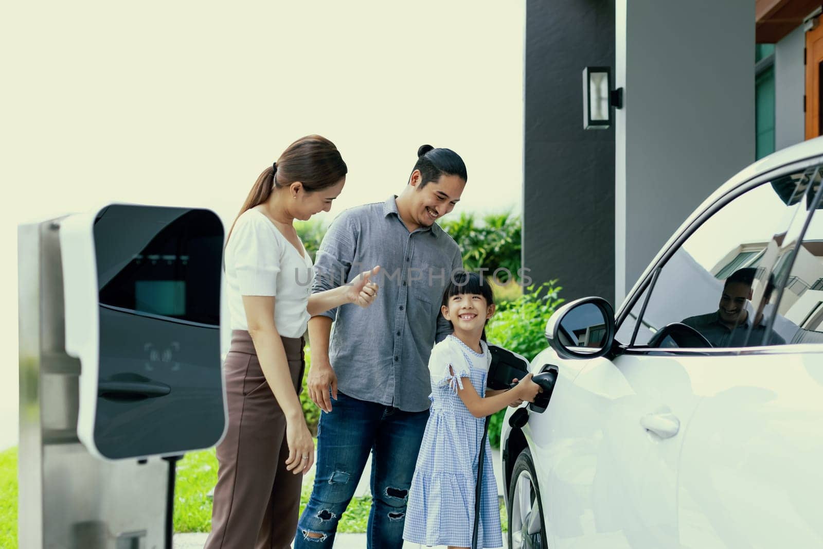 Progressive young parent teach daughter how to recharge or refuel EV car at home charging station. Green and clean energy from electric vehicle for healthy environment. Eco power from renewable source