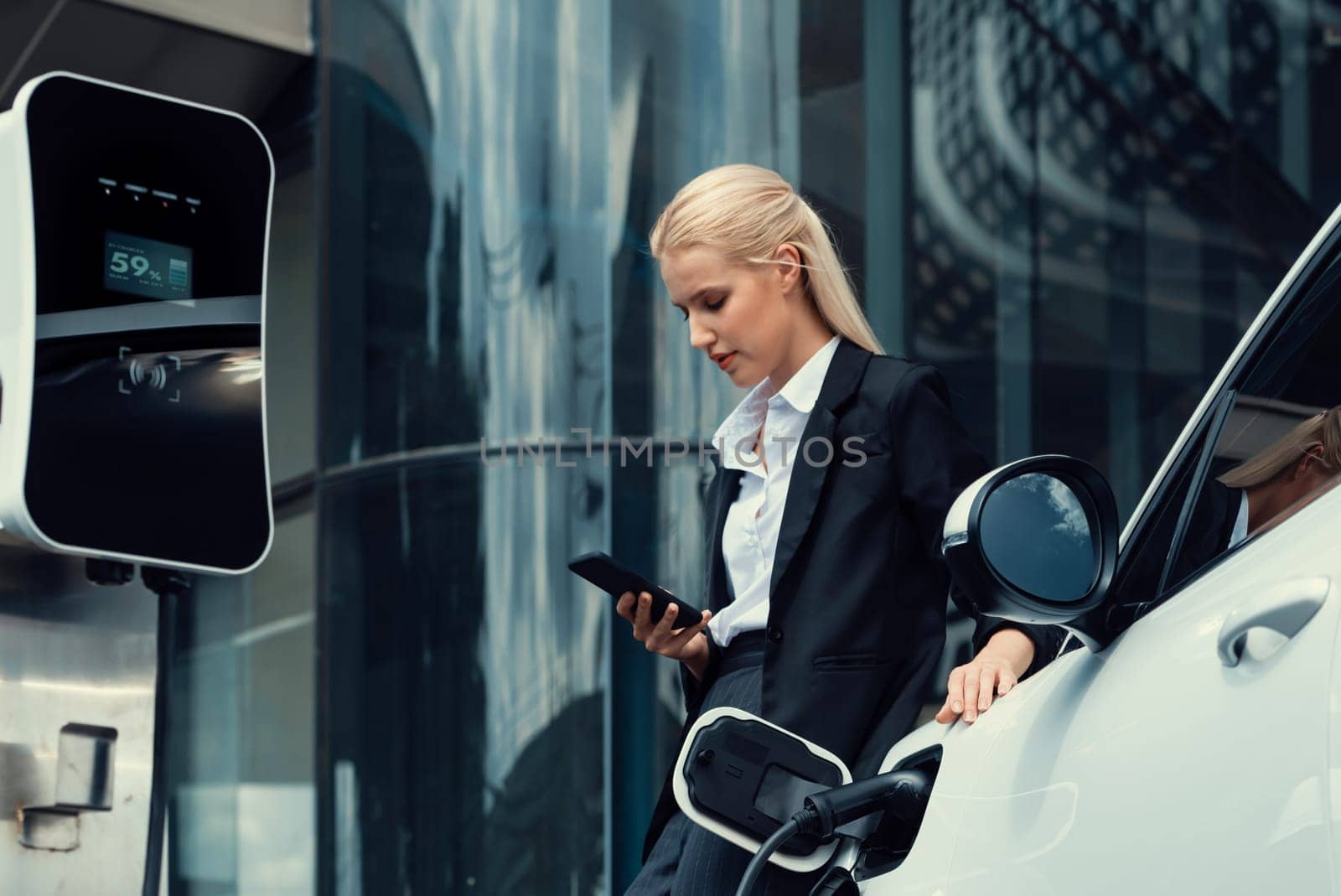 Businesswoman wearing black suit using smartphone, leaning on electric car recharge battery at charging station in city residential building with condos and apartment. Progressive lifestyle concept.