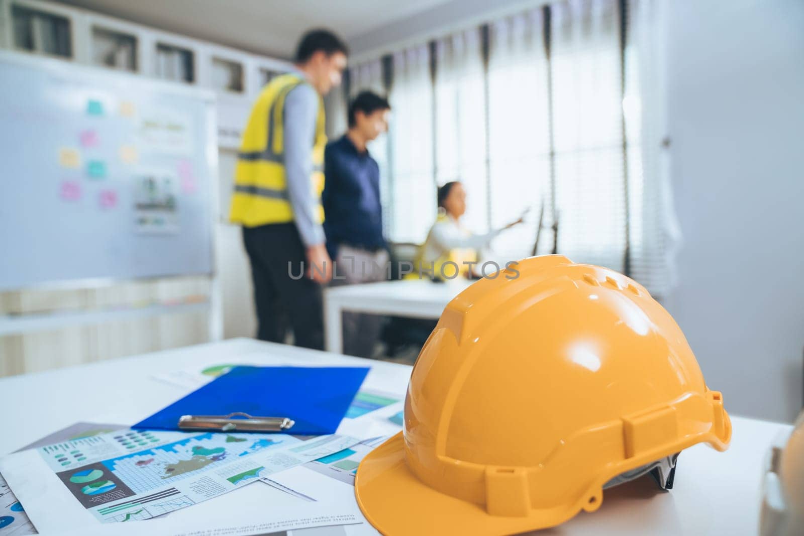 Concept of engineering equipments of protective helmet for competent engineer on piles of paper works. Blurred image of engineers and employer working together at the desk.