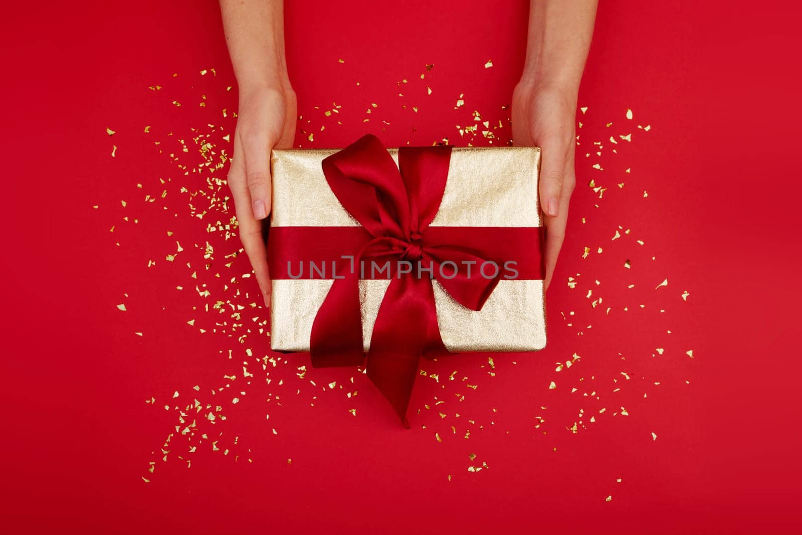 Hands give a gold gift box with a red bow on isolated burgundy background top view