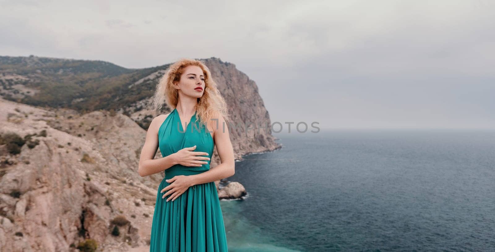 Side view a Young beautiful sensual woman in a mint long dress posing on a volcanic rock high above the sea during sunset. Girl on the nature on overcast sky background. Fashion photo