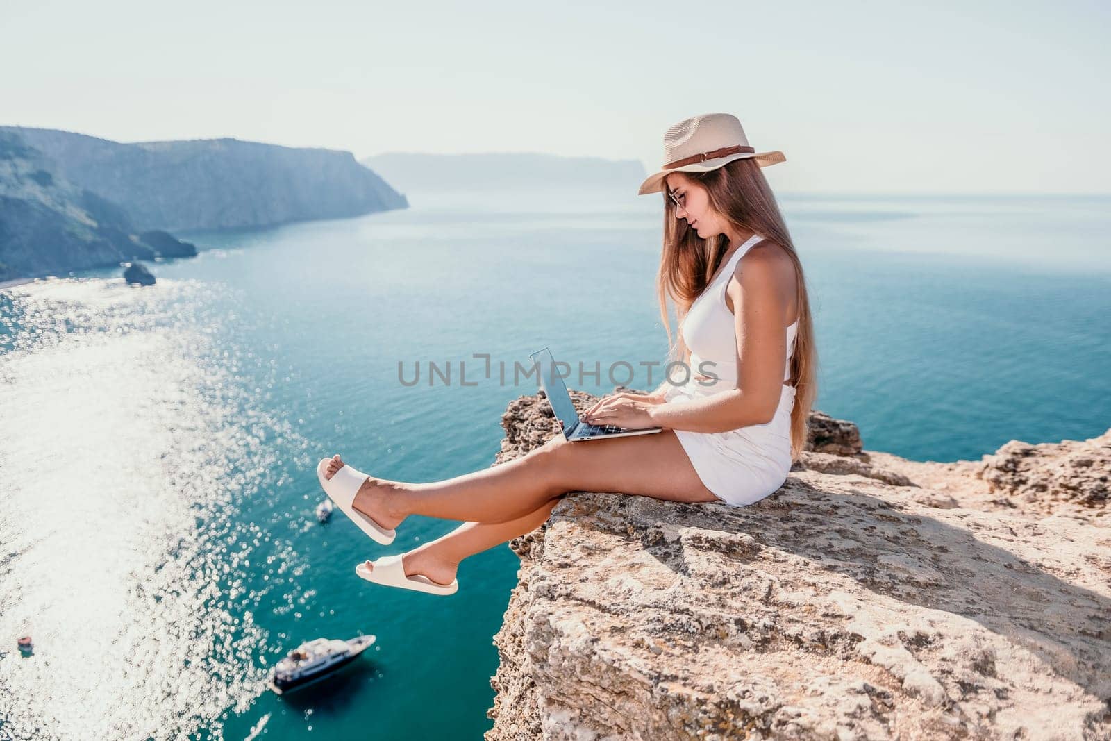 Successful business woman in yellow hat working on laptop by the sea. Pretty lady typing on computer at summer day outdoors. Freelance, travel and holidays concept.