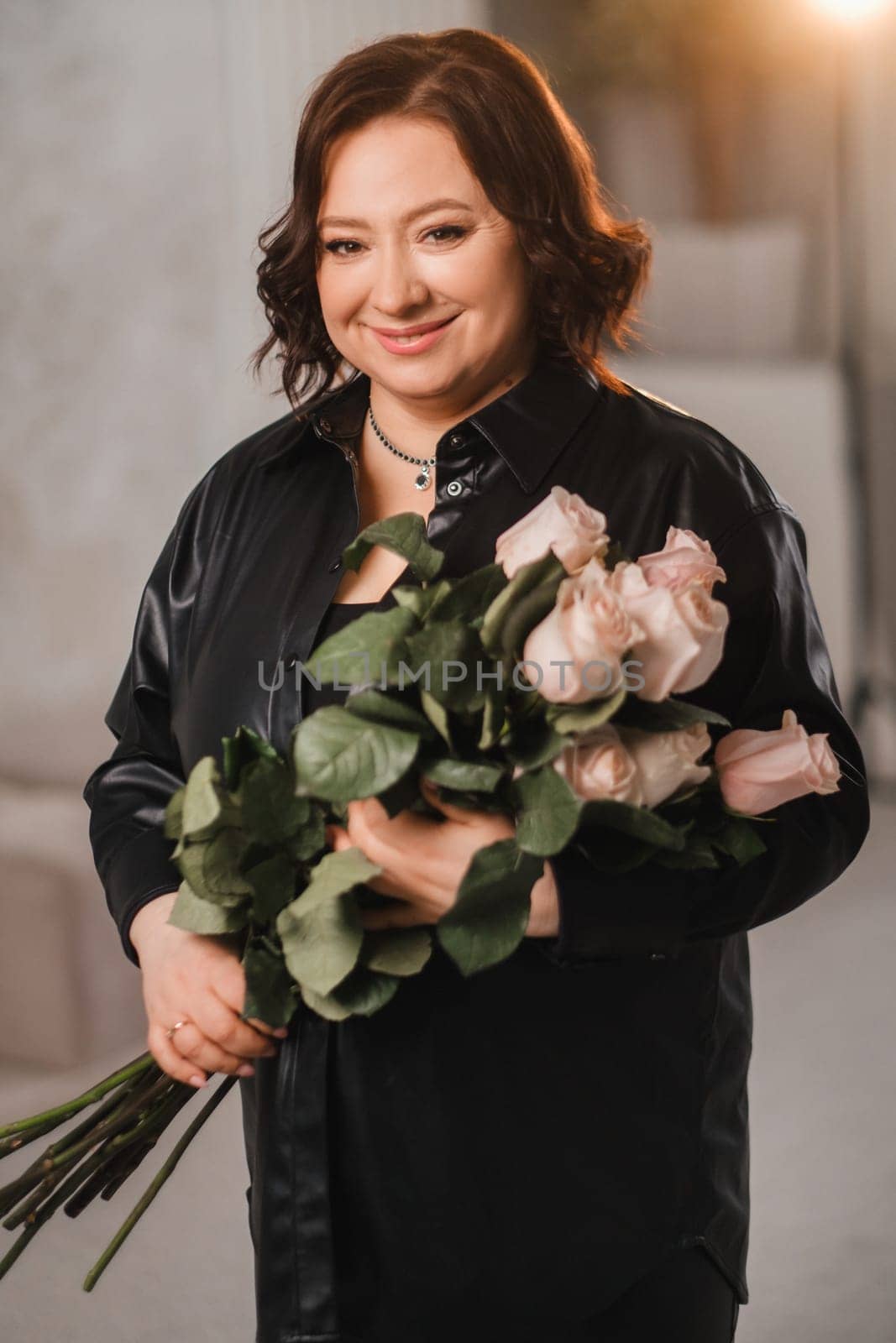 a stylish adult woman in black leather clothes stands with a bouquet of pink roses in the interior.
