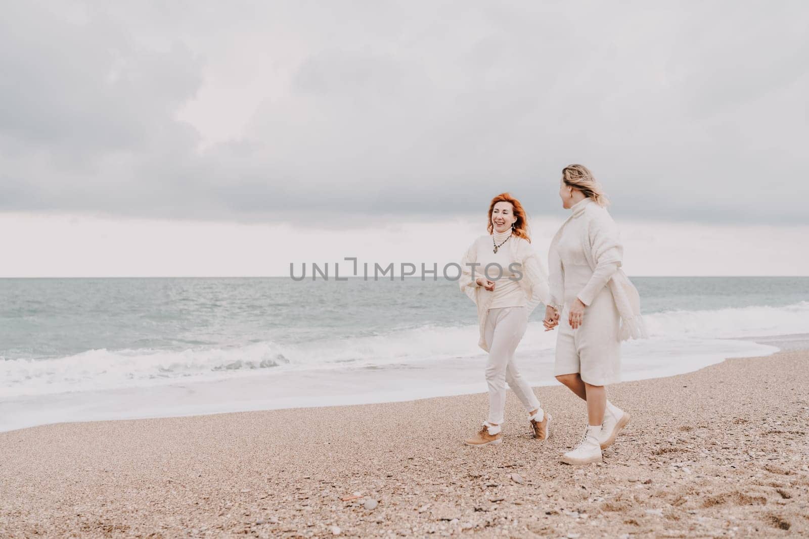 Women sea walk friendship spring. Two girlfriends, redhead and blonde, middle-aged walk along the sandy beach of the sea, dressed in white clothes. Against the backdrop of a cloudy sky and the winter sea. Weekend concept. by Matiunina