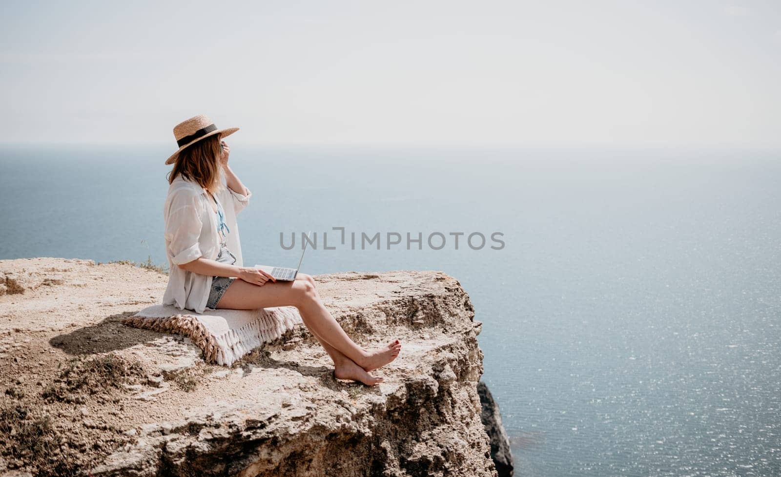 Successful business woman in yellow hat working on laptop by the sea. Pretty lady typing on computer at summer day outdoors. Freelance, travel and holidays concept.