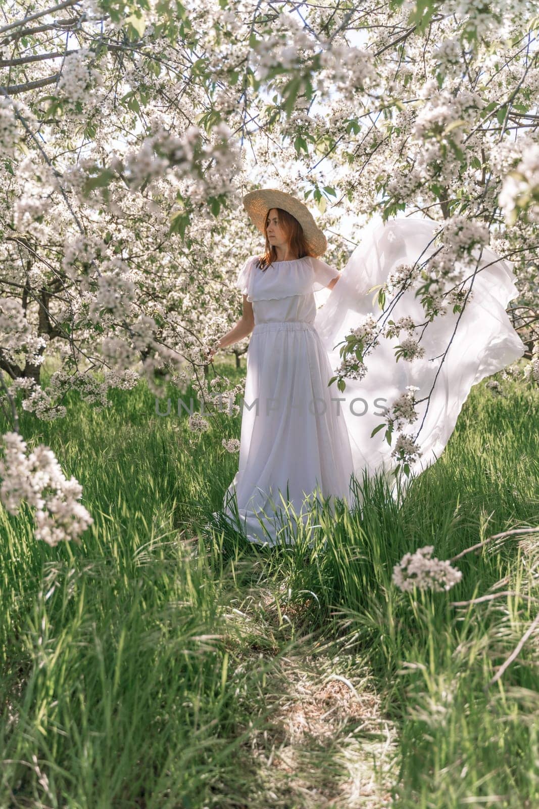 Woman cherry orchard. A happy woman in a long white long dress walks through the green spring blooming cherry garden. Happy cheerful princess bride. The fabric of the skirt flutters in the wind. by Matiunina