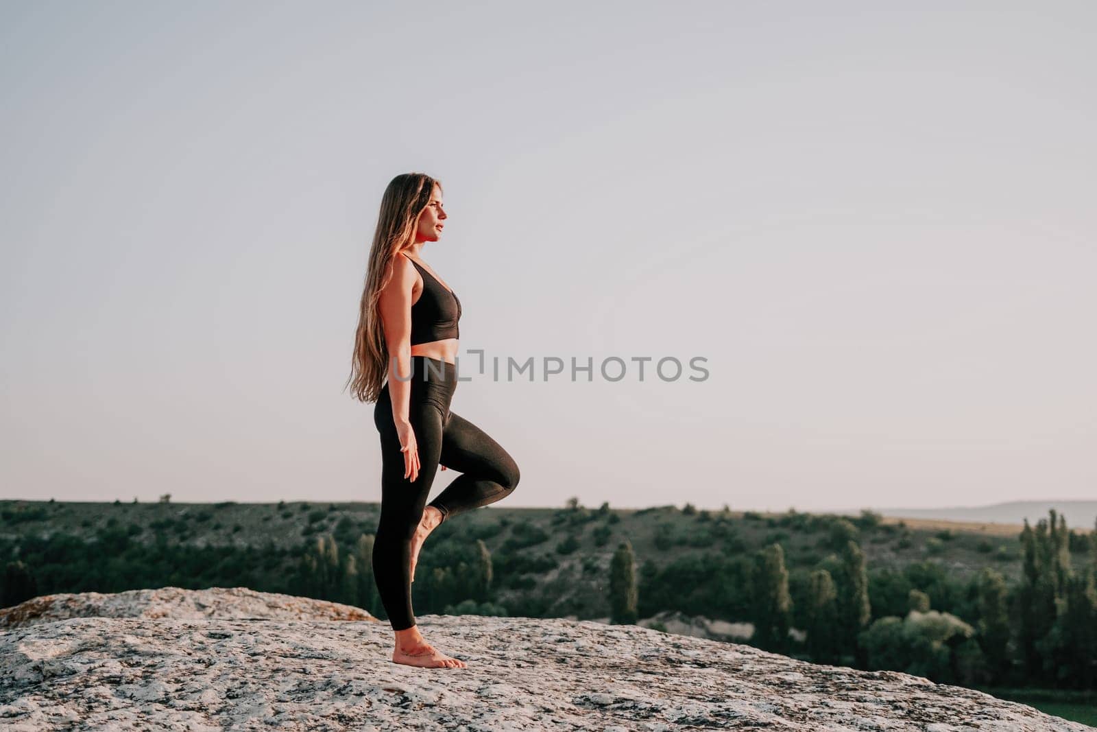 Well looking middle aged woman with long hair, fitness instructor in leggings and tops doing stretching and pilates on the rock near forest. Female fitness yoga routine concept. Healthy lifestyle.