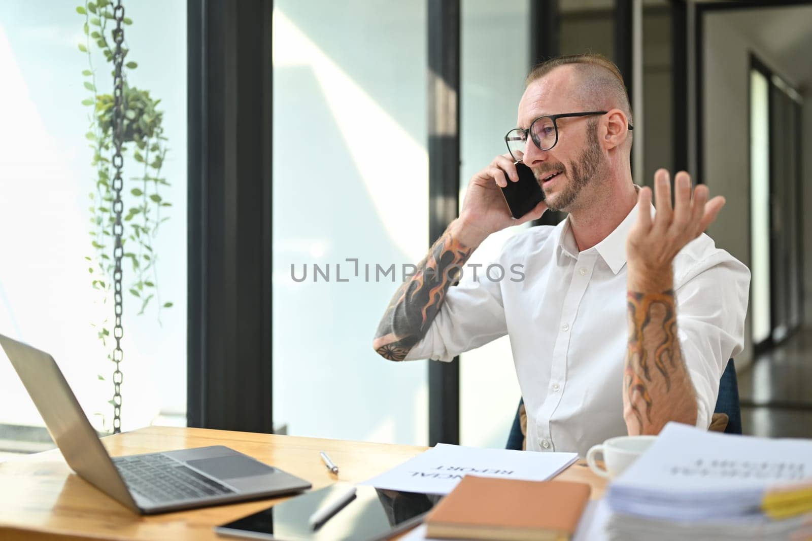 Confident male manager wearing glasses and white shirt talking on phone at office desk with laptop.