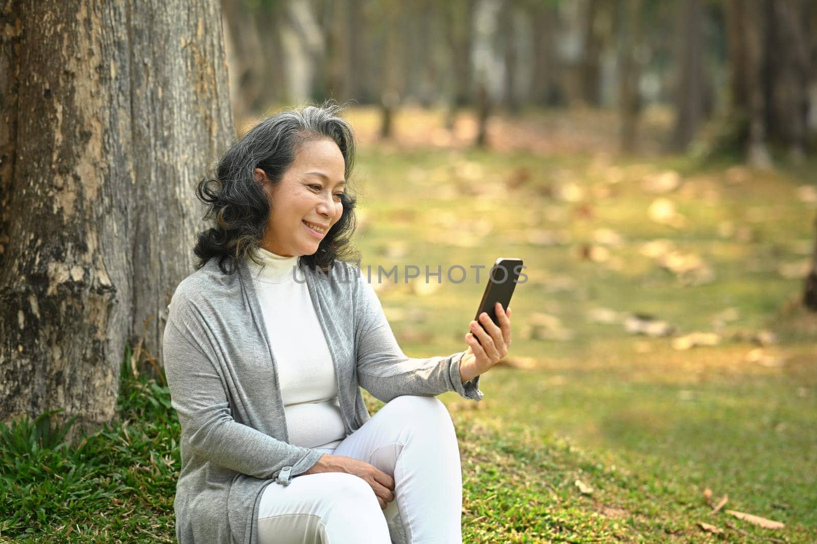 Positive middle aged woman using smart phone under tree on green grass at sunny beautiful day by prathanchorruangsak