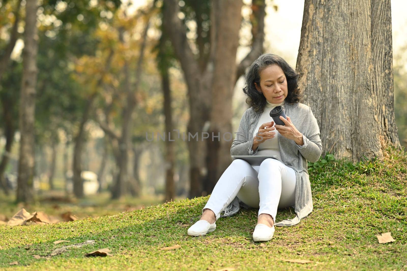Pleasant middle age woman drinking coffee, relaxing under a tree on beautiful garden background by prathanchorruangsak