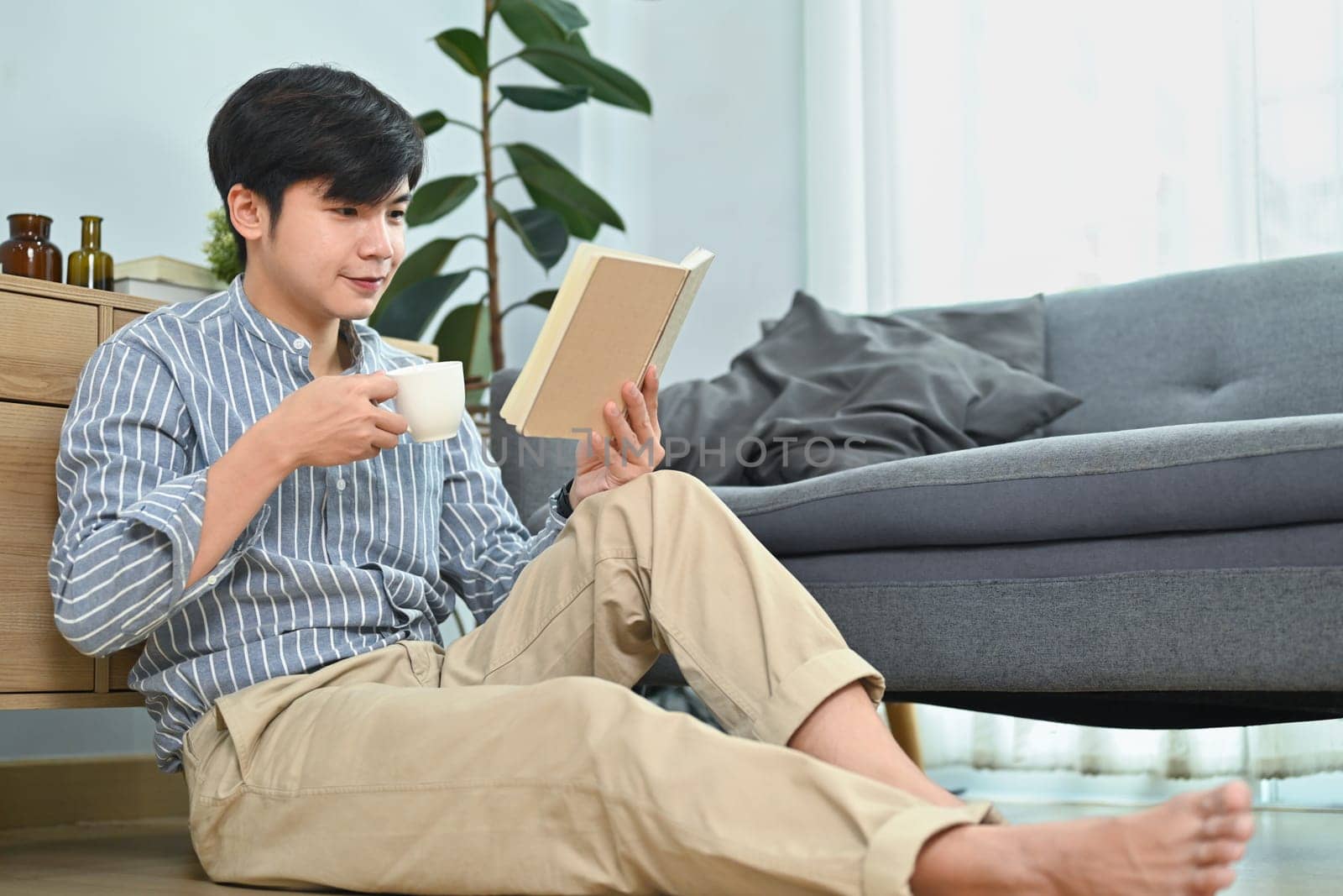 Satisfied millennial man drinking hot coffee and reading book in bright living room. Leisure and people concept by prathanchorruangsak