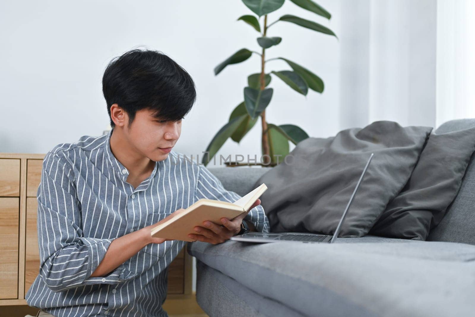Pleasant asian man sitting on floor and reading book, spending leisure time at home. Leisure and people concept.