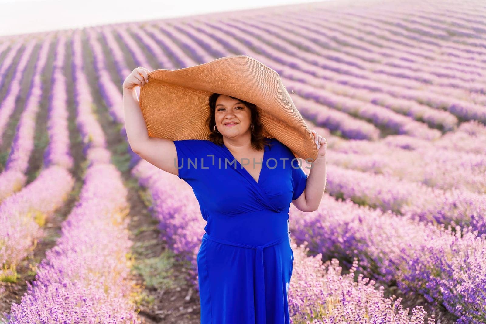 Woman lavender field sunset. Romantic woman walks through the lavender fields. illuminated by sunset sunlight. She is wearing a blue dress with a hat