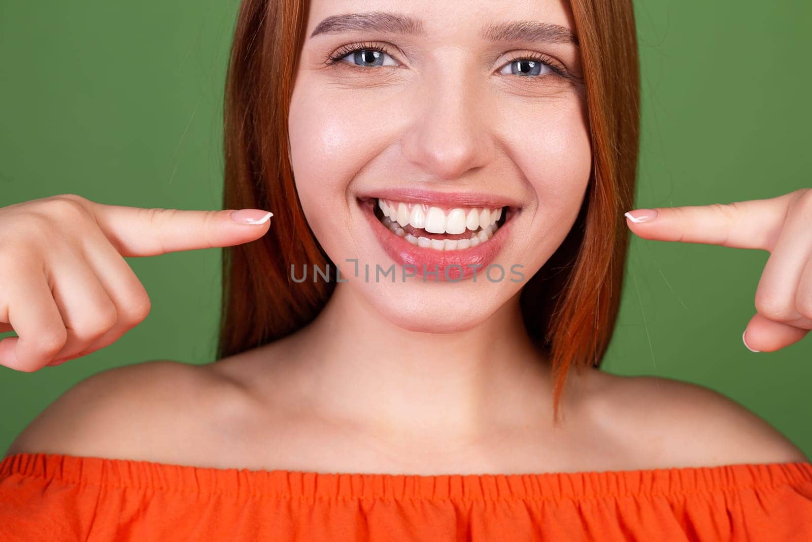 Young red hair woman in casual orange blouse on green background point fingers on white teeth perfect smile by kroshka_nastya