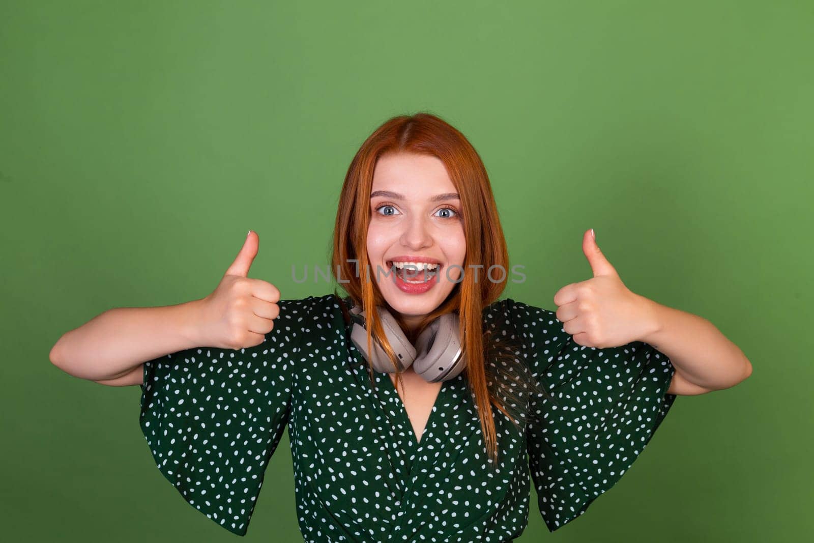 Young red hair woman on green background with wireless headphones happy laugh shows thumbs up by kroshka_nastya