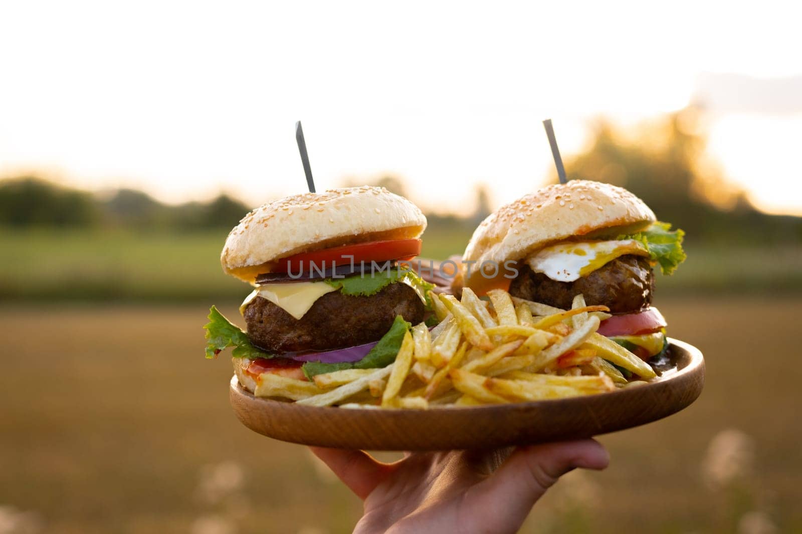 Two hamburgers with beef burger cutlet, fried onion, ketchup sauce and cheese in traditional buns, served on wood chopping board. A hand holds a tray in the open air of the sunset. High quality photo
