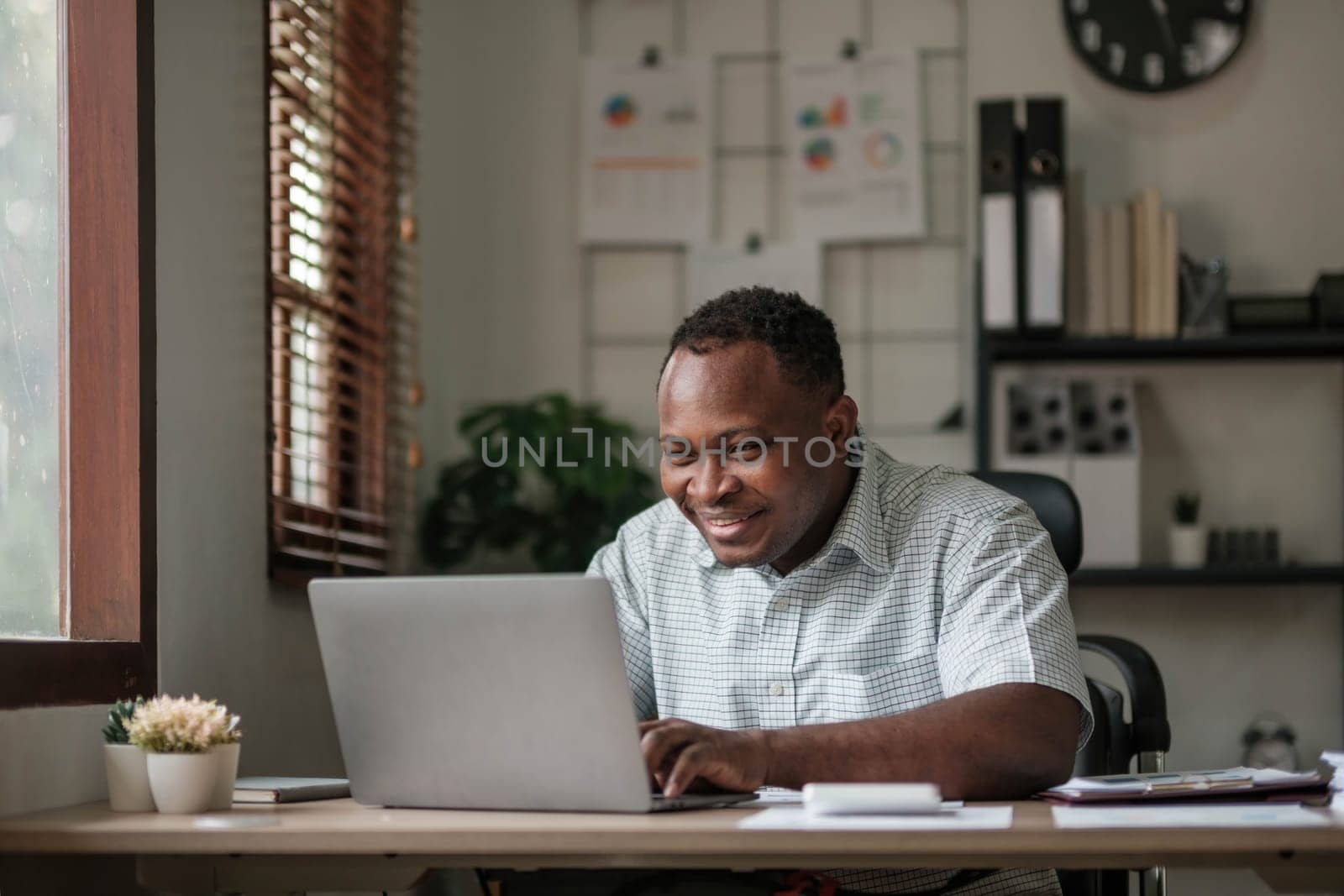 Smiling black man using laptop at home in living room. Happy mature businessman send email and working at home. African american freelancer typing on computer with paperworks and documents on table...