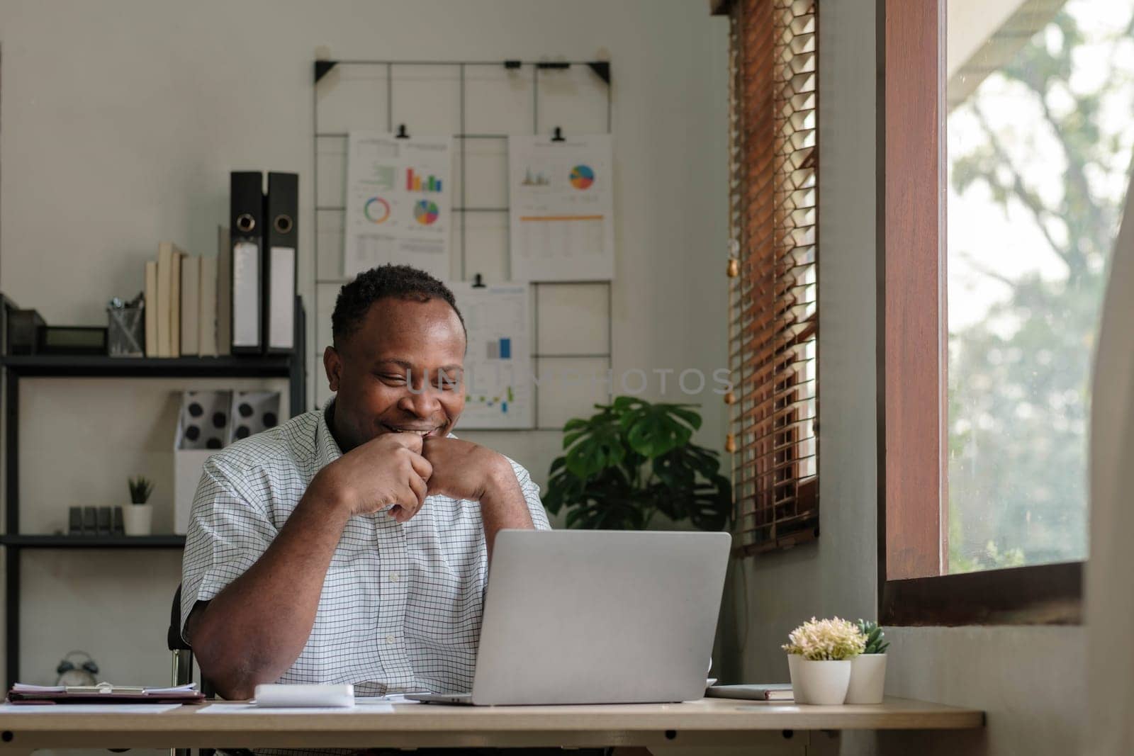 Smiling black man using laptop at home in living room. Happy mature businessman send email and working at home. African american freelancer typing on computer with paperworks and documents on table. by wichayada