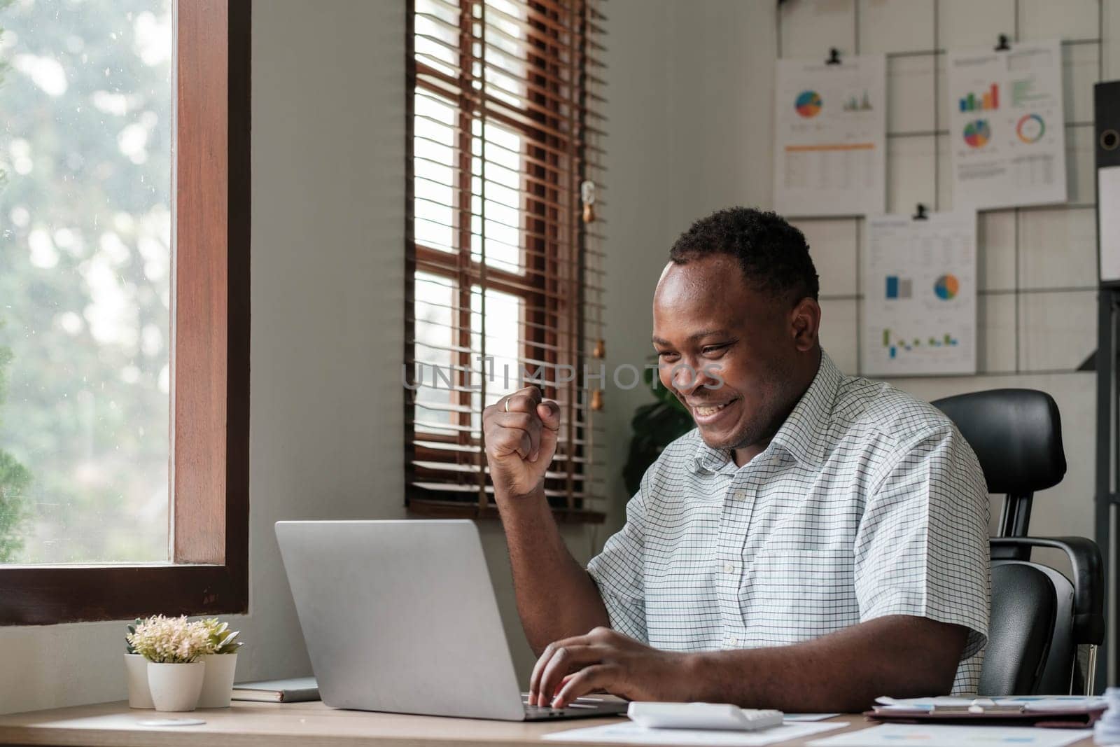 Smiling black man using laptop at home in living room. Happy mature businessman send email and working at home. African american freelancer typing on computer with paperworks and documents on table. by wichayada