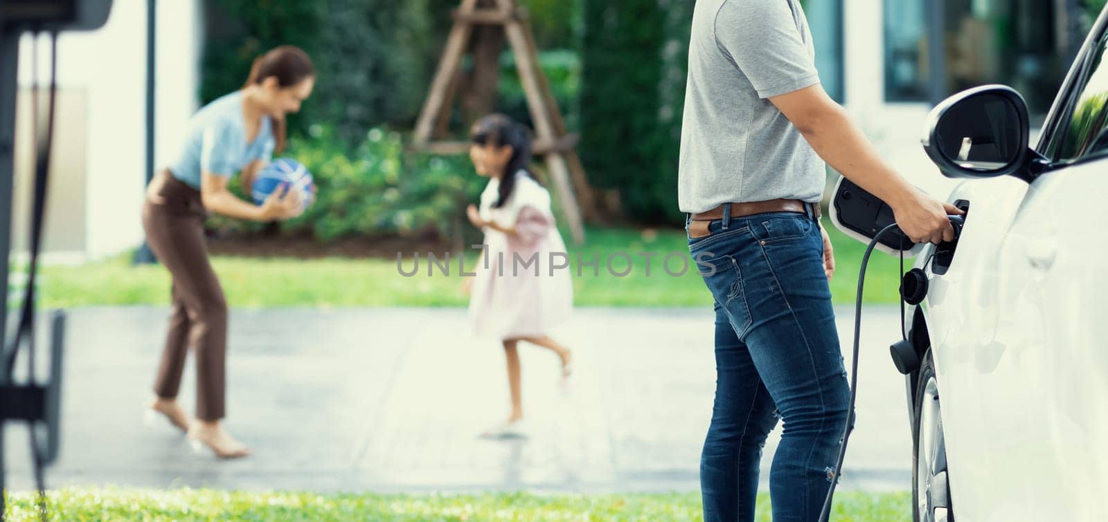 Focus image of progressive man charging electric car from home charging station with blur mother and daughter playing together in the background.