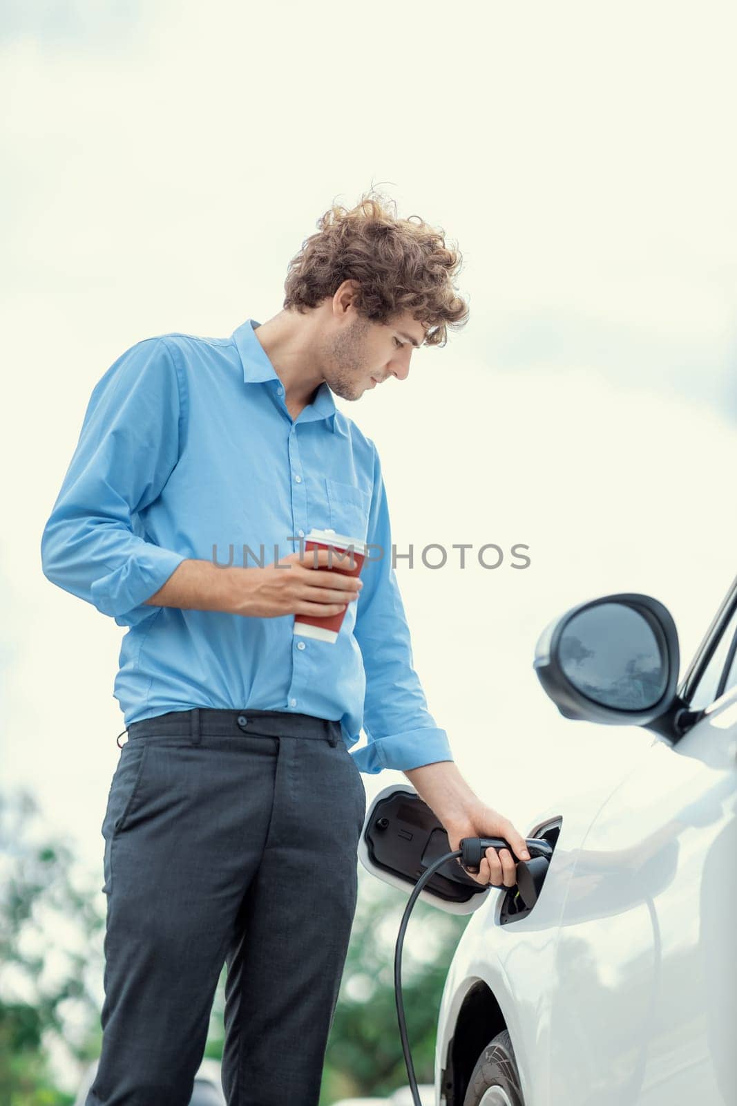 Progressive eco-friendly concept of parking EV car at public electric-powered charging station in city with blur background of businessman leaning on recharging-electric vehicle with coffee.