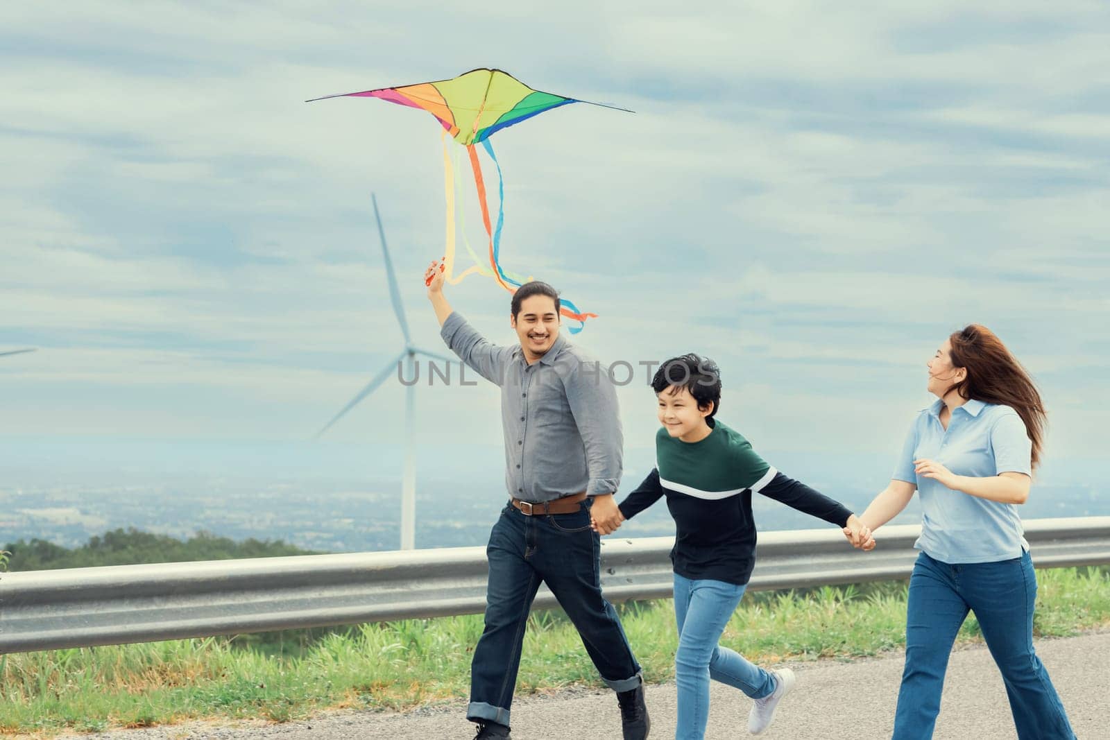 Progressive happy carefree family vacation concept. Young parents mother father and son run along and flying kite together on road with natural scenic on mountain and wind turbine background.