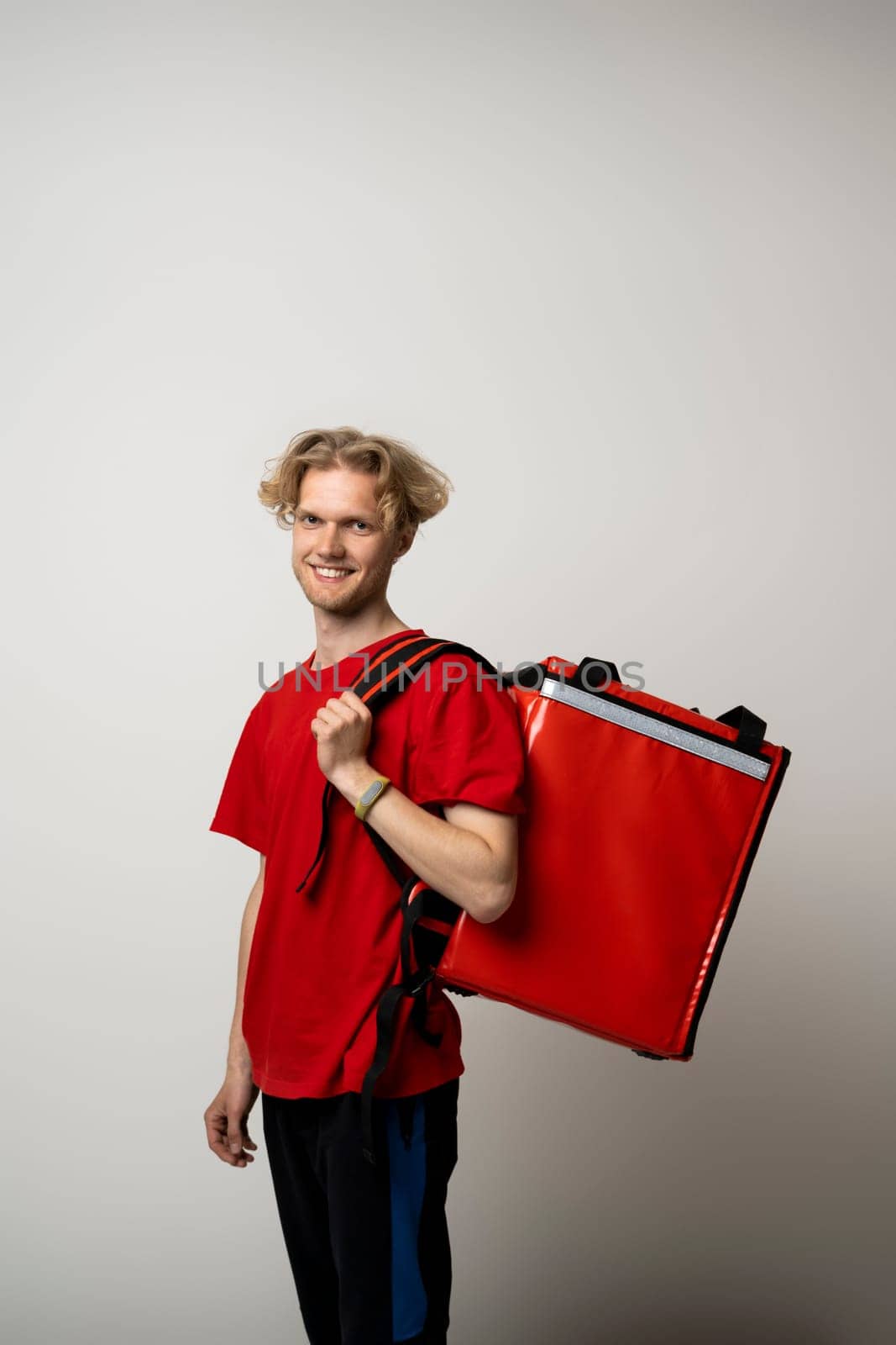 Smiling young courier with curly hair in red uniform with red thermo bag on white background. Food delivery service. by vovsht