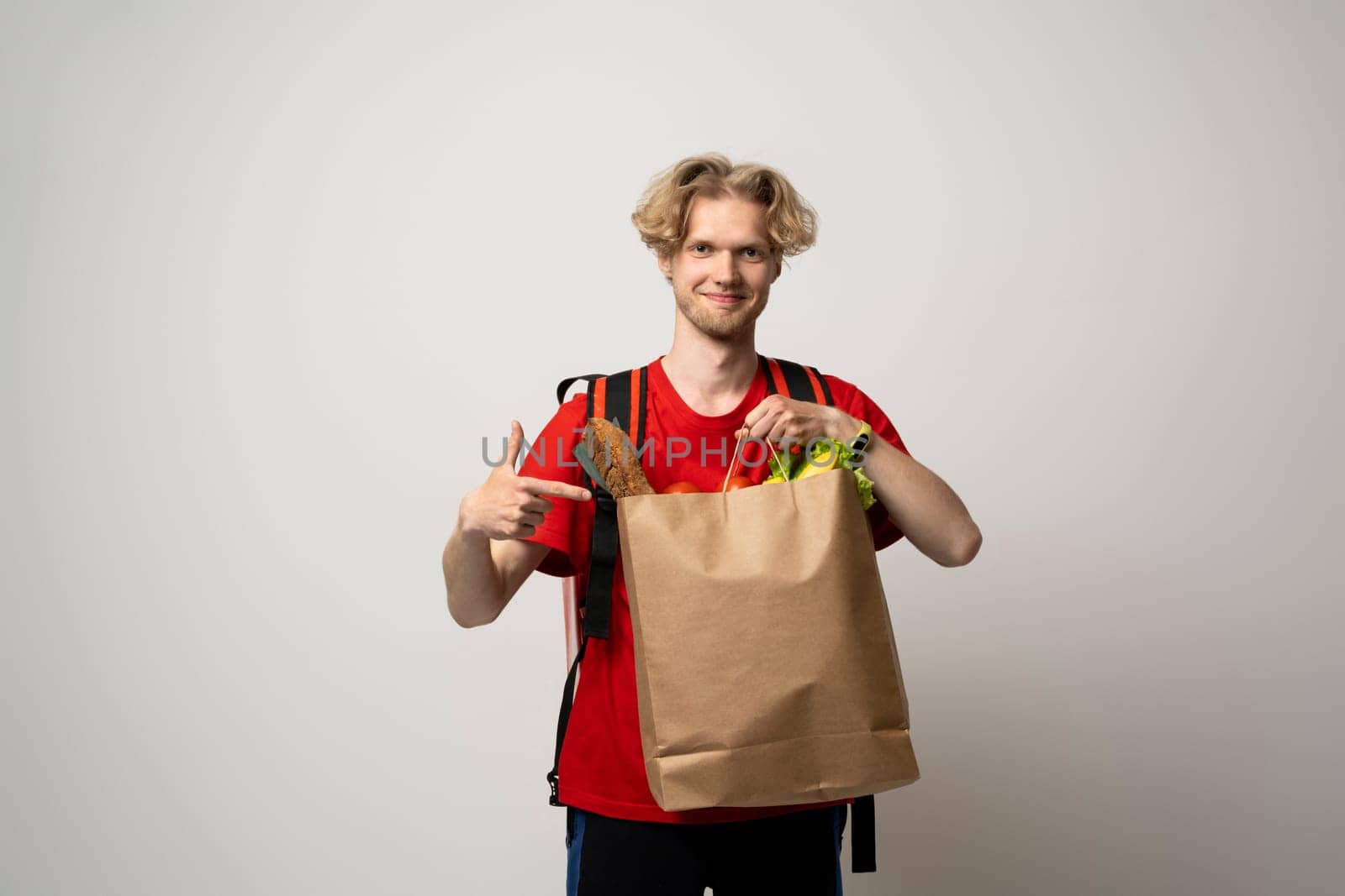 Food delivery service. Portrait of pleased delivery man in red uniform smiling while carrying paper bag with food products isolated over white background