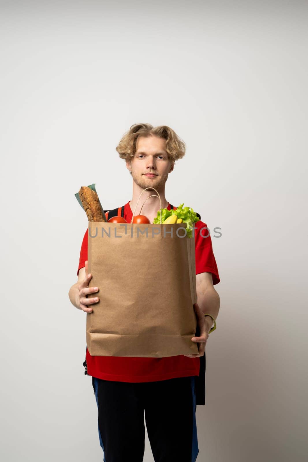 Portrait of pleased delivery man in red uniform smiling while carrying paper bag with food products isolated over white background