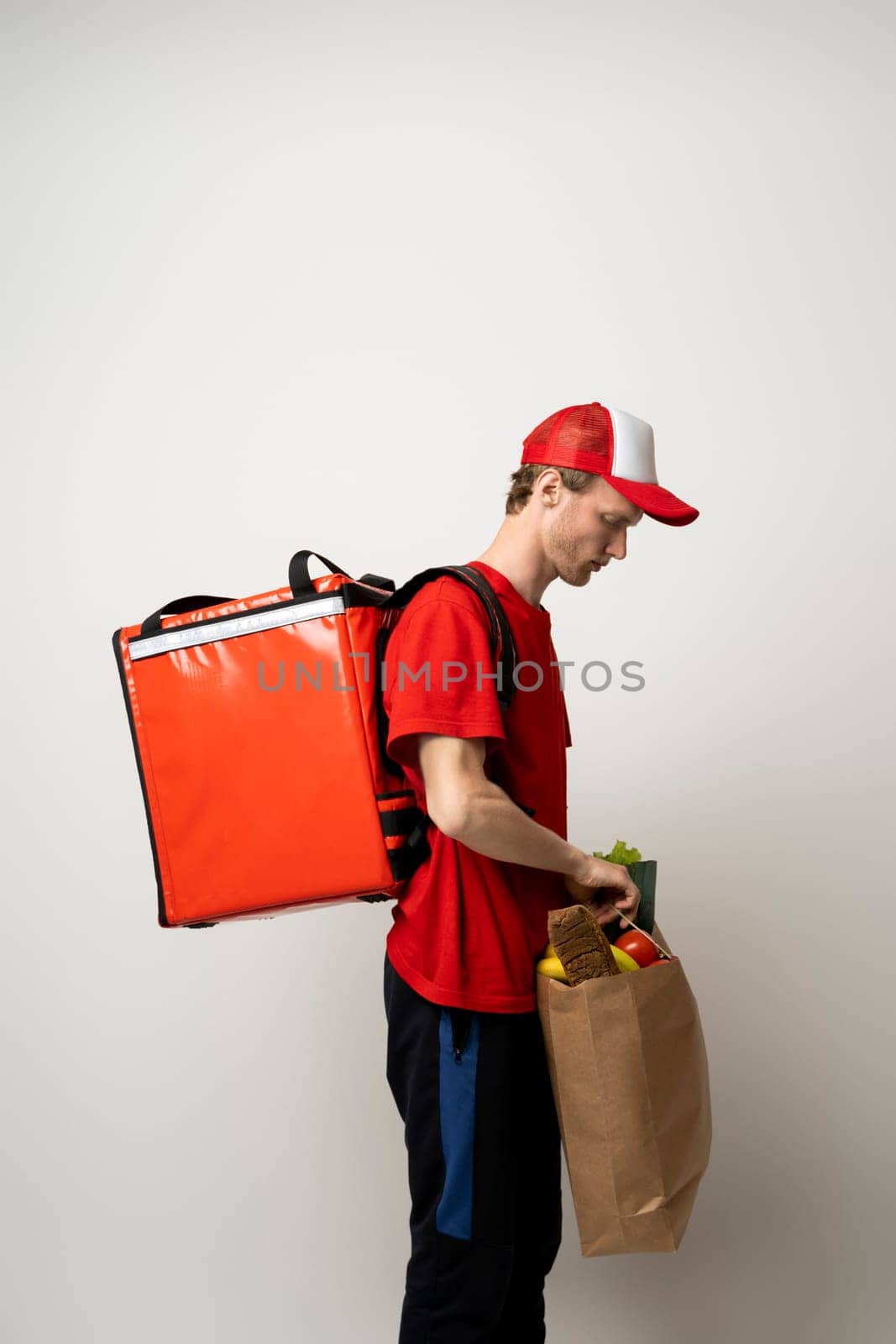 Young man in a red t-shirt and a cap holding paper bag with products on white background. Food delivery service