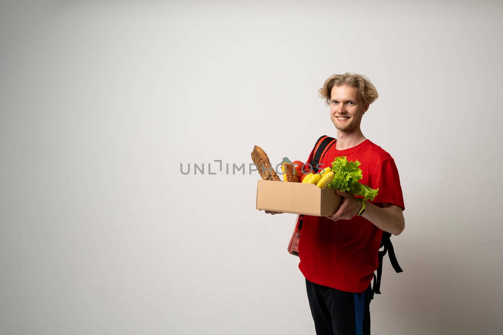 Happy delivery man in red uniform with a box full of groceries over white background. Express delivery, food delivery, online shopping concept