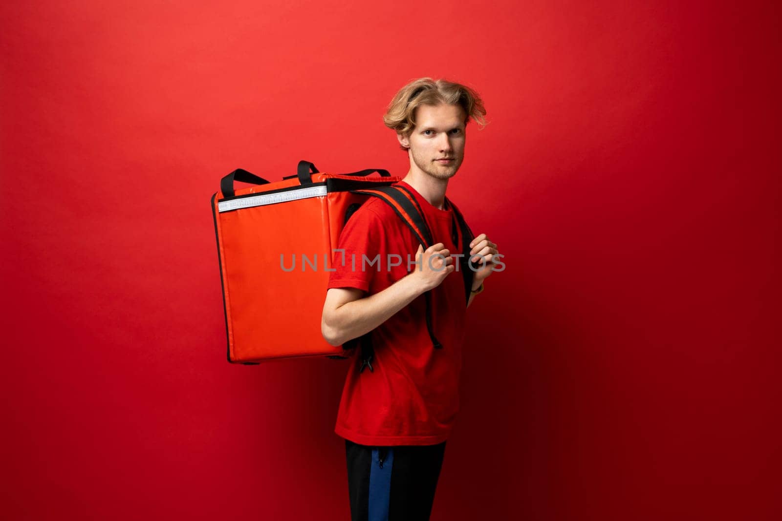 Best Delivery Service Concept. Portrait of confident smiling male courier wearing red uniform and thermo backpack bag looking at camera isolated on red background