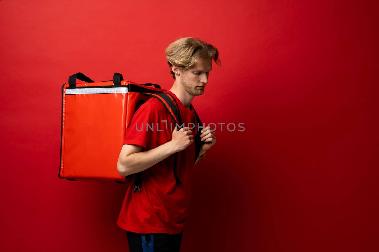 Side view of young courier with curly hair in red uniform with red thermo bag on red background. Food delivery service